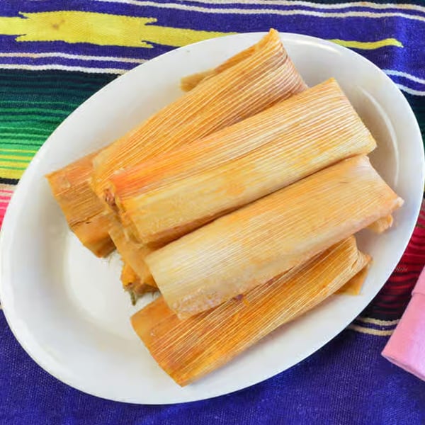 A white oval plate filled with several golden-brown tamales, resting on a colorful striped Mexican blanket.