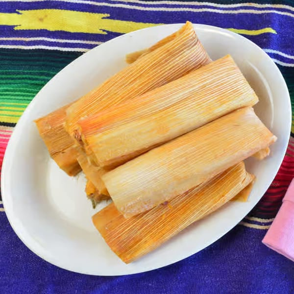 A white oval plate filled with several golden-brown tamales, resting on a colorful striped Mexican blanket.