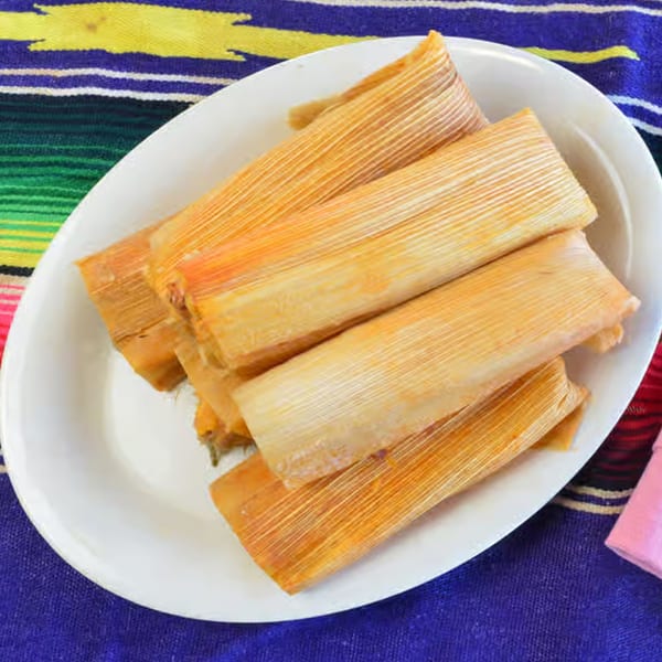 A white oval plate holds several golden tamales wrapped in corn husks, on a colorful striped tablecloth.